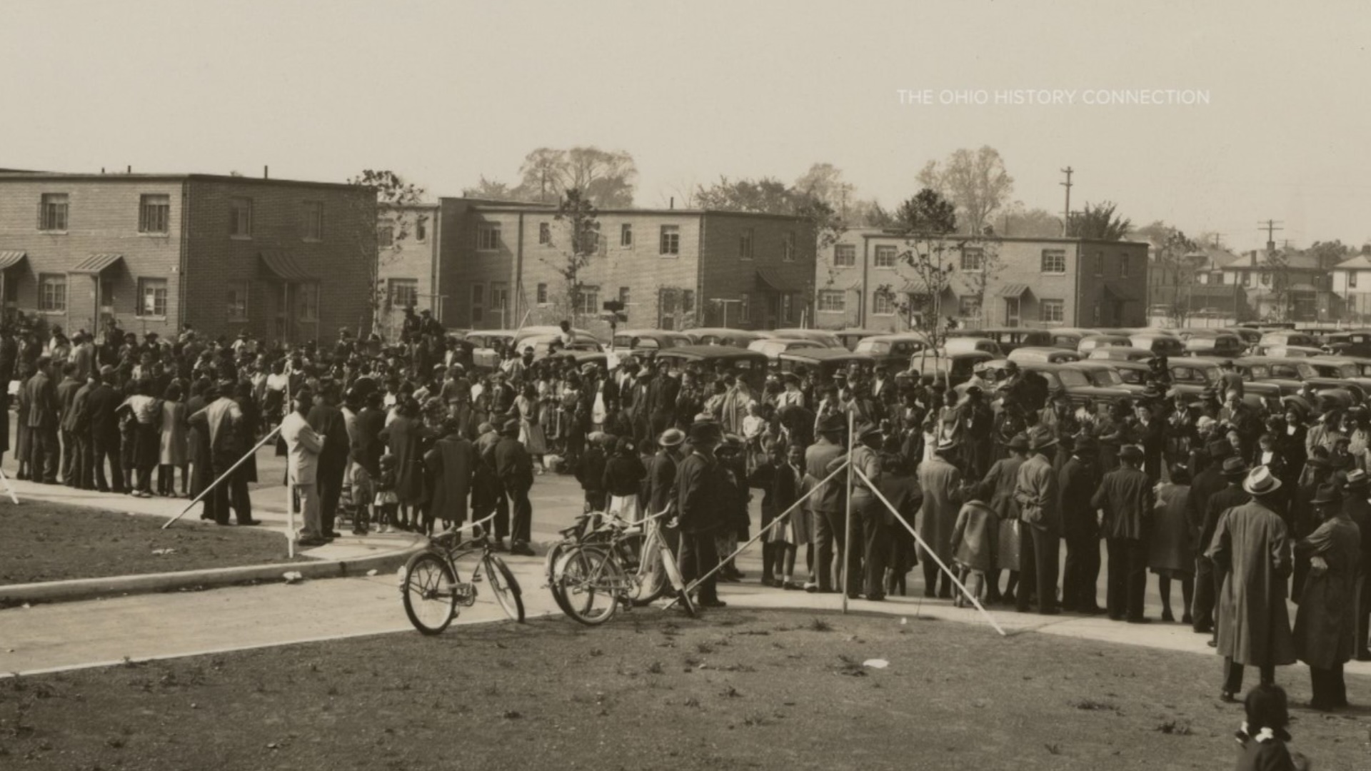 A crowd gathers in front of the original Poindexter Village, with two bikes sitting on the grass behind the crowd, which faces the complex’s buildings. The image is an old black-and-white photograph.