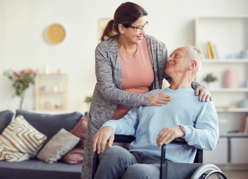A middle-aged white man sits in a wheelchair in a living room. A middle-aged woman stands next to him with her arms resting on his shoulders.