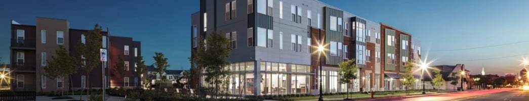 A three-story apartment building with a lower-level community space is lit up at dusk.
  The building sits on a street corner and streetlights shine around the building, sprinkled amid several young trees.