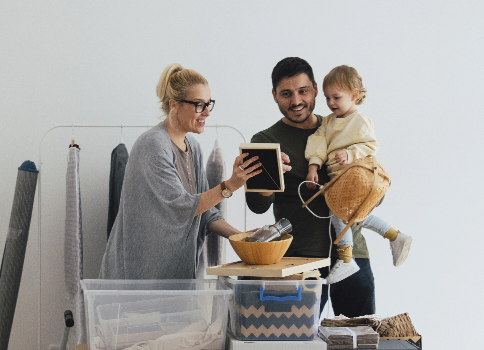 A multiracial three-person family of a man, woman and small child unpack boxes and smile at a framed photo.