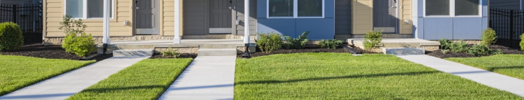 The sun shines on the fresh-cut lawn of the front of a house with gray and blue siding. Landscaping sits below the two front windows of the house.