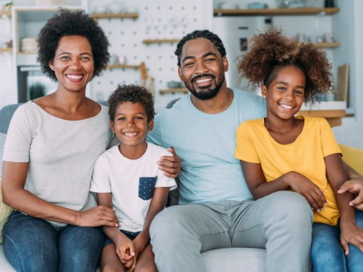 An African American family of four sits smiling on their couch in their home. It
consists of a woman, man, young girl and a slightly younger boy.