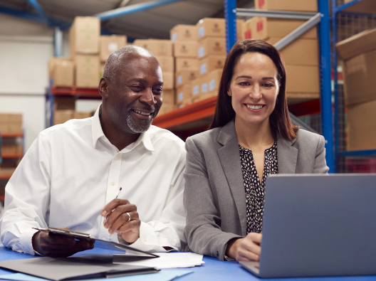 A middle-aged African American businessman and middle-aged businesswoman look at a laptop screen together. They are sitting in a warehouse with boxes stacked on shelves behind them.